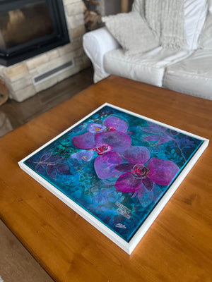 A framed floral artwork featuring large purple orchids over a vivid blue and teal background, displayed on a wooden table in a living room with a white sofa and fireplace visible.