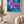 A framed floral wall art print hangs on a beige wall in a styled living room. The artwork shows large purple orchid flowers with pink centers over a vivid blue and teal textured background.