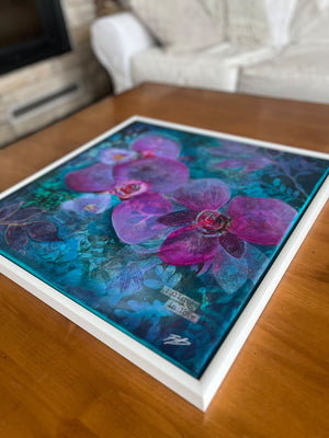 A framed floral art print featuring large purple orchids over a vivid blue and teal background, displayed on a wooden table in a living room setting.