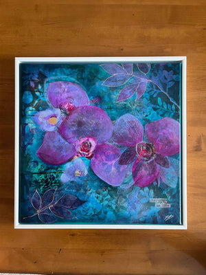 A framed mixed-media painting of purple orchids and blue leaves on a textured teal background, displayed on a wooden surface.