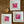 Three white square framed art prints with pink botanical abstract designs, arranged on a brown wooden floor next to a small wooden heart-shaped object.