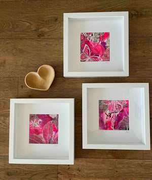 Three white square framed art prints with pink botanical abstract designs, arranged on a brown wooden floor next to a small wooden heart-shaped object.