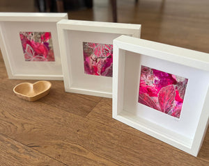 Three white square shadow box frames with vibrant pink and red botanical leaf prints, arranged on a wooden floor next to a small wooden heart-shaped dish.
