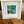 A framed tropical art print featuring green palm leaves and pink flowers on a turquoise background, matted in white and housed in a white square frame, displayed on a wooden table next to a wooden heart-shaped bowl.