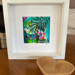 A framed tropical art print featuring green palm leaves and pink flowers on a turquoise background, matted in white and housed in a white square frame, displayed on a wooden table next to a wooden heart-shaped bowl.