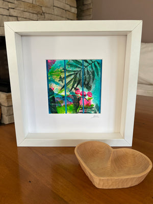 A framed tropical art print featuring green palm leaves and pink flowers on a turquoise background, matted in white and housed in a white square frame, displayed on a wooden table next to a wooden heart-shaped bowl.