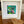 A white square frame containing a colorful abstract tropical artwork with green leaves and pink flowers, displayed on a wooden table next to a small wooden bowl.