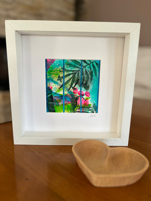 A white square frame containing a colorful abstract tropical artwork with green leaves and pink flowers, displayed on a wooden table next to a small wooden bowl.