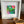 A white square picture frame with a white mat holding a small colorful abstract artwork, displayed on a wooden surface next to a shallow wooden bowl.