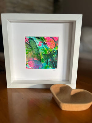 A white square picture frame with a white mat holding a small colorful abstract artwork, displayed on a wooden surface next to a shallow wooden bowl.