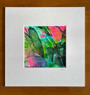 A square abstract botanical art print with vibrant tropical leaves in green, pink, and blue, matted on a wide white sheet of paper and photographed on a wooden surface.