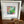 A white square shadow box frame containing a colorful abstract painting with green, pink, and gold brushstrokes, displayed on a wooden surface next to a small wooden heart-shaped object.