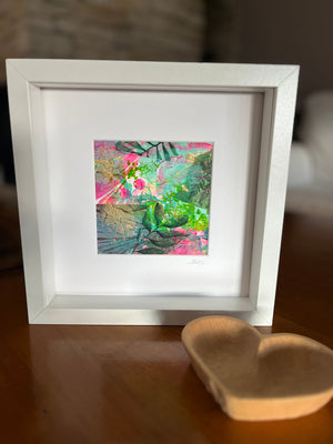 A white square shadow box frame containing a colorful abstract painting with green, pink, and gold brushstrokes, displayed on a wooden surface next to a small wooden heart-shaped object.