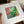A hand holds a matted art print featuring a colorful abstract botanical painting with green leaves, pink flowers, and gold leaf details on a white mat board.