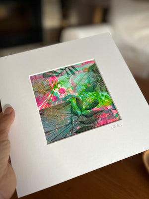 A hand holds a matted art print featuring a colorful abstract botanical painting with green leaves, pink flowers, and gold leaf details on a white mat board.