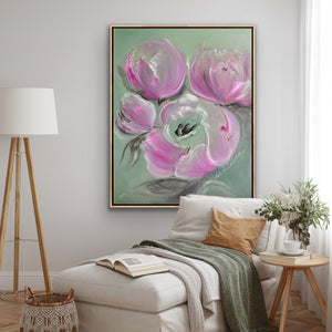 A framed floral wall art print featuring large pink flowers on a soft mint green background, displayed above a light beige sofa in a styled living room with a floor lamp, pillow, blanket, and side table.