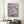 A framed canvas painting of pink peony flowers on a sage green background, displayed on a light beige wall in a modern living room setting with a gray pouf, black coffee table, and white curtains.
