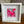 A square white shadow box frame containing a vibrant pink and red abstract art print with a butterfly motif, displayed on a wooden table.