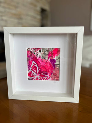 A square white shadow box frame containing a vibrant pink and red abstract art print with a butterfly motif, displayed on a wooden table.