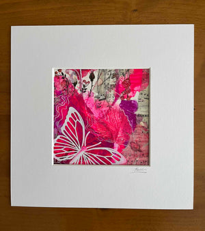 A square mixed-media art print featuring a vibrant pink and red butterfly design over a background of pink paint splatters, musical notes, and vintage postcard textures. The print is mounted on a wide white mat board, which is placed on a wooden surface.