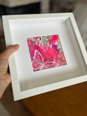 A hand holds a white square shadow box frame containing a colorful abstract art print with a prominent pink butterfly design, set against a blurred indoor background.