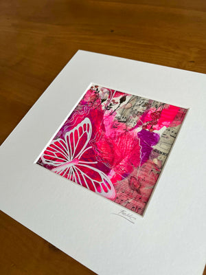 A square mixed-media art print featuring vibrant pink, red, and white butterfly and floral designs over a sheet music background, presented with a wide white mat board on a wooden table.