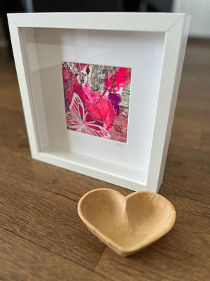 A white square shadow box frame displaying a vibrant pink and red butterfly art print, styled on a wooden tabletop next to a small wooden heart-shaped dish.
