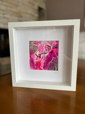 A white square framed art print with a white mat, featuring an abstract pink and magenta botanical design, displayed on a wooden table.