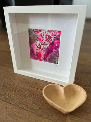 A white shadow box frame containing a vibrant pink and red abstract art print, displayed on a wooden table next to a small wooden heart-shaped bowl.