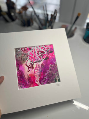 A matted art print held by a hand, featuring a vibrant pink and magenta abstract botanical design with white leaf patterns and dark red accents, set against a blurred background of paintbrushes.