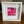 A white square shadowbox frame containing a vibrant abstract floral art print in shades of hot pink, magenta, and red, displayed on a wooden table in front of a stone fireplace.
