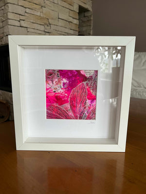 A white square shadowbox frame containing a vibrant abstract floral art print in shades of hot pink, magenta, and red, displayed on a wooden table in front of a stone fireplace.