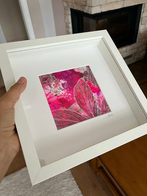 A hand holds a white square shadowbox frame containing a vibrant pink and magenta abstract floral artwork with metallic gold leaf details, set against a blurred home interior background.