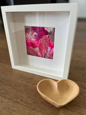 A white square shadow box frame displaying a vibrant pink and red abstract leaf artwork, styled on a wooden tabletop next to a small light wood heart-shaped dish.
