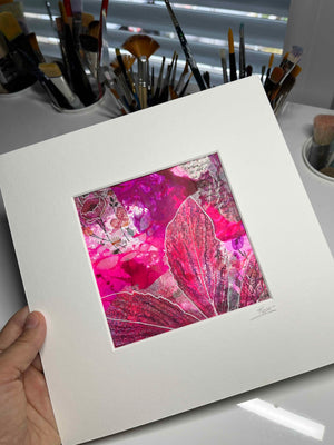 A hand holds a matted art print featuring a vibrant abstract floral design in shades of magenta, pink, and red with silver glitter accents. The background shows a white desk with various paintbrushes in cups.
