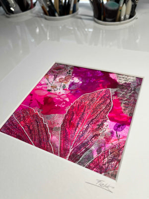 A matted art print featuring a vibrant abstract botanical design in shades of magenta, pink, and red, displayed on a glossy white surface with paintbrushes in the background.