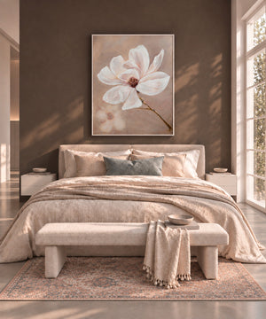 A styled bedroom interior with a large framed floral wall art piece above a bed. The artwork shows a white magnolia flower with a soft beige background.